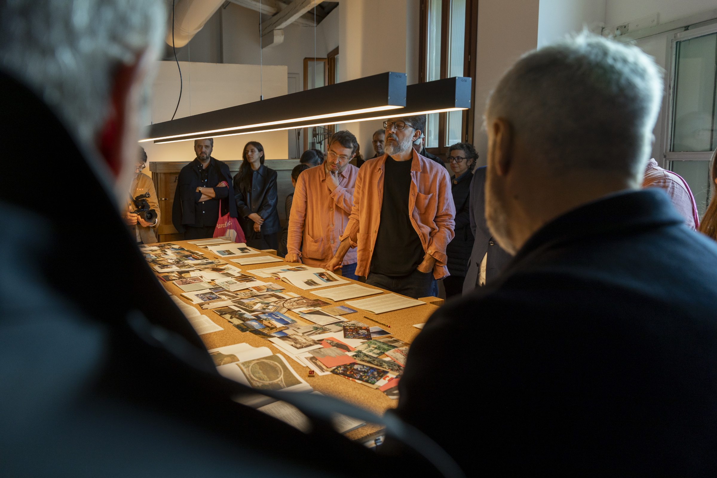 Grupo de pessoas reunido em torno de uma mesa iluminada, observando fotografias e documentos expostos numa sala de exposição.
